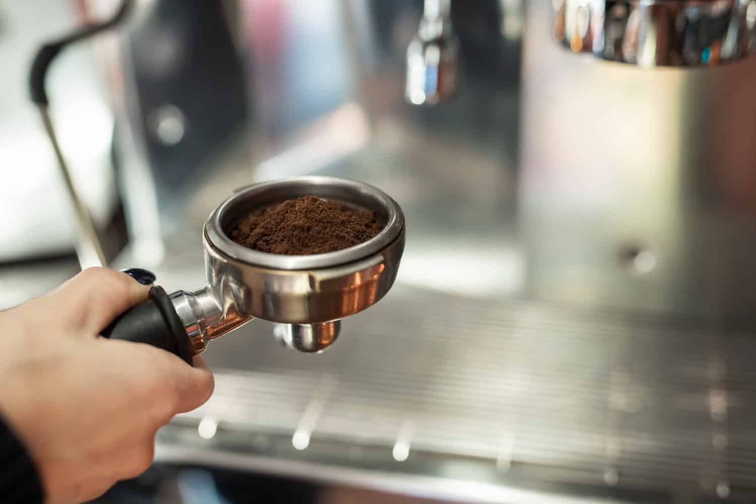 Mano che prepara un portafiltro riempito di caffè macinato davanti a una macchina da caffè professionale.