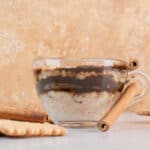 Tazza di vetro con marocchino caffè, decorata con bastoncini di cannella, circondata da biscotti su una superficie chiara con sfondo beige.