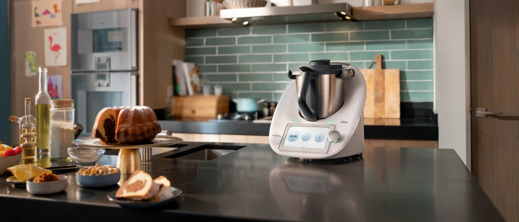 Robot da cucina Bimby TM6 posizionato su un piano di lavoro con un dolce fatto in casa, ingredienti freschi e utensili da cucina in una cucina moderna con sfondo di piastrelle verdi.