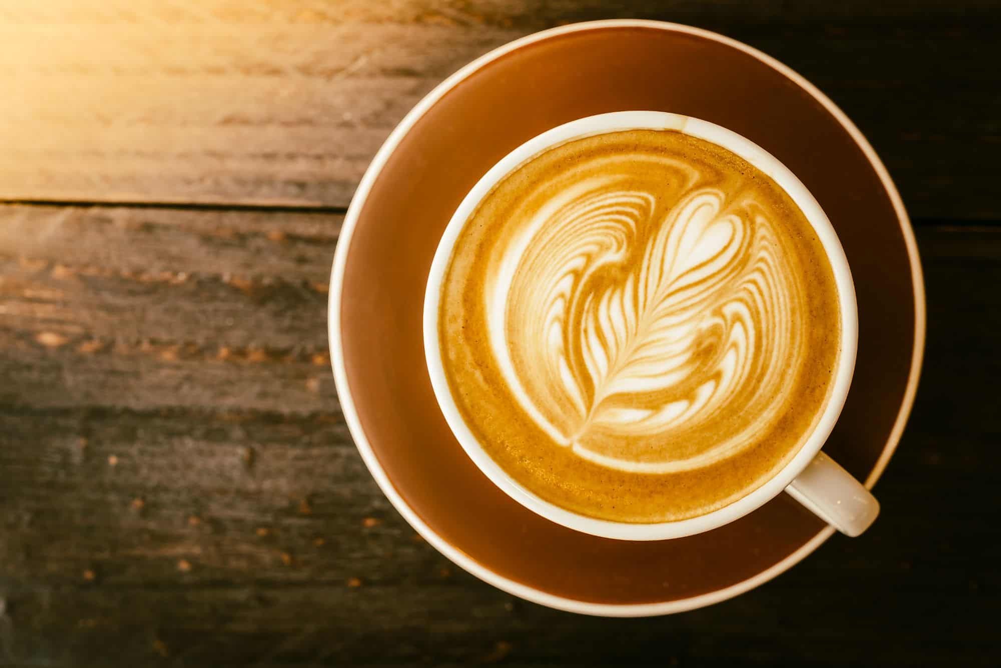 Tazza di cappuccino con decorazione di latte art a forma di foglia, vista dall'alto, su piattino marrone e tavolo in legno scuro.